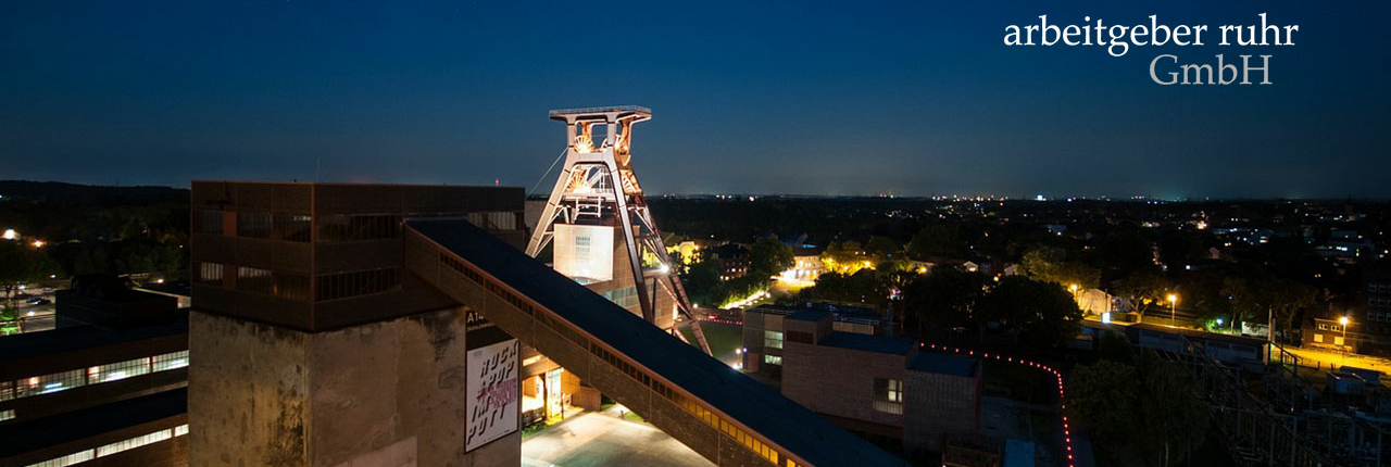 Blick auf Essen bei Nacht, im Vordergrund der beleuchtete Förderturm der Zeche Zollverein Logo der arbeitgeber ruhr Gmbh